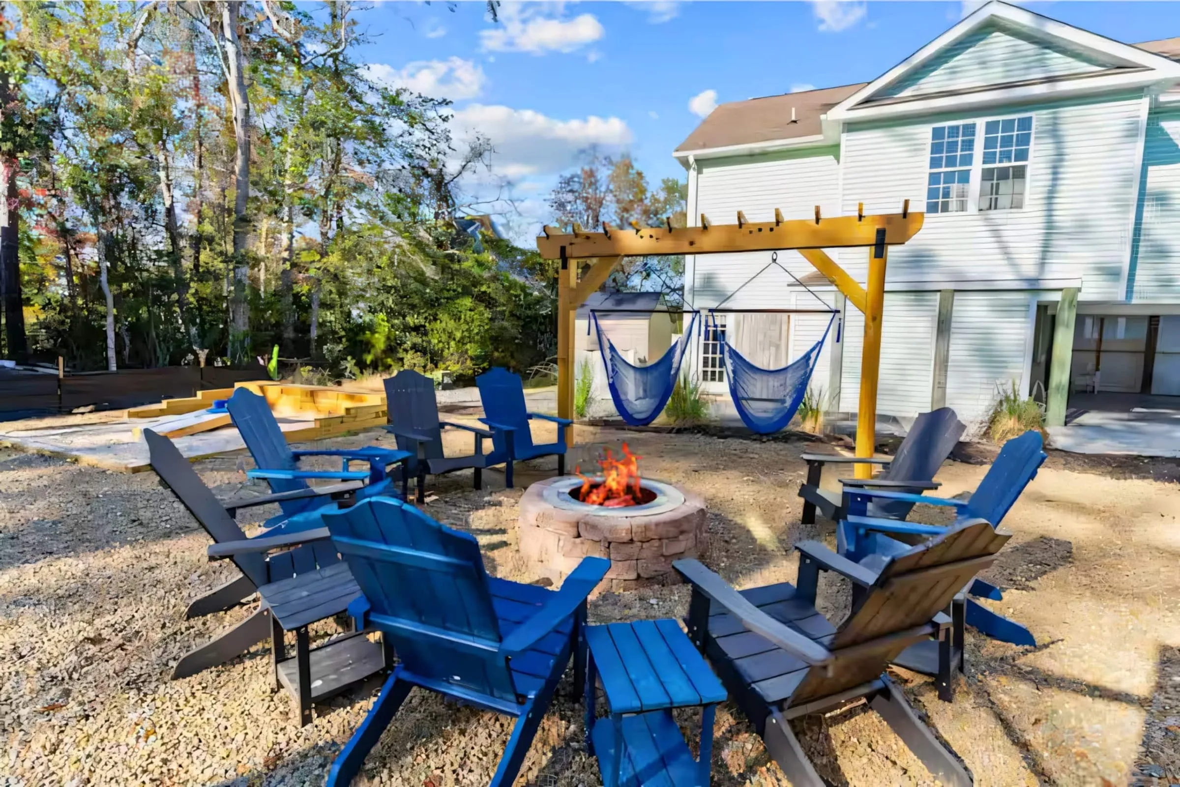 Blue Crab Beach House at dusk: hammocks under the pergola, Adirondack chairs around a lit fire pit, the blue cottage glowing behind